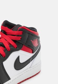 Středně vysoké tenisky z černé kůže s červenými akcenty, bílými panely, texturovanou gumovou podrážkou a červenými tkaničkami. Logo na kotníku.