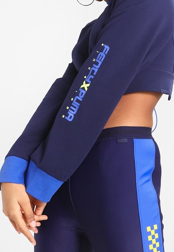 Granatowa krótka bluza i dopasowane granatowe legginsy z niebieskimi akcentami. Bluza ma logo „FENTY x PUMA” w żółtym i zielonym kolorze na rękawie.