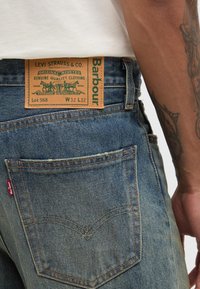 Calças de ganga com um tom azul claro. Apresenta um remendo em couro castanho com letras verdes, design com rebites, costura laranja e um logótipo na bolsa traseira.