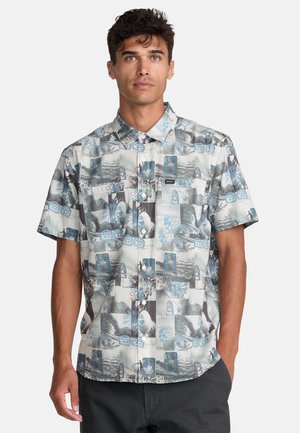 Uomo che indossa una camicia a maniche corte con bottoni, con motivo astratto e floreale blu e grigio, in piedi con una mano in tasca su sfondo bianco.