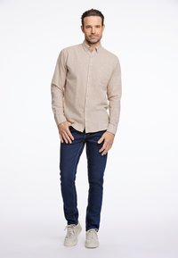 Camisa beige de botones con un bolsillo, combinada con vaqueros oscuros y zapatillas claras. La camiseta cuenta con un tejido suave y texturizado.