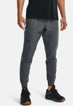 Mężczyzna w szarych spodniach dresowych Under Armour i czarnych butach sportowych stoi z rękami swobodnie opuszczonymi wzdłuż ciała.