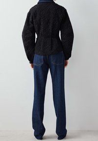 Svart quiltad jacka med strukturerad tyg, ståkrage och figursydd midja. Parat med högmidjade blå denimjeans.