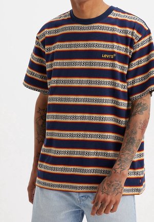 Homme portant un t-shirt Levi's rayé aux motifs bleu, rouge et beige, avec des tatouages sur les avant-bras et un jean bleu clair, sur un fond uni.