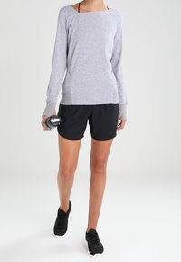 Ljusgrå långärmad topp med ribbade muddar, ihop med svarta shorts och svarta sneakers. Modellen håller en vattenflaska.