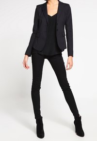 Svart skräddarsydd kavaj med notch-lapeller, bärs över en svart topp; matchas med slim fit svarta byxor och svarta ankelstövlar. Slät tyg med struktur.