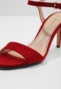 Sandale à talon haut en daim rouge avec bride à la cheville et boucle, semelle intérieure beige avec étiquette de la marque Esprit.