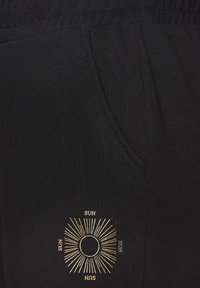 Schwarze Sweatpants aus weichem Material, ausgestattet mit einer Seitentasche und einer goldenen Grafik einer Sonne mit Strahlen und dem Wort "SUN", das darum wiederholt wird.
