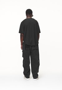 Uomo di spalle che indossa una maglietta nera oversize, pantaloni cargo neri con una toppa e sneakers bianche, in piedi su uno sfondo bianco.