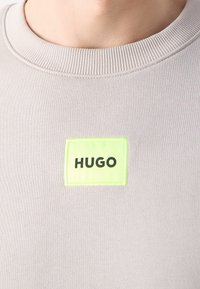 Detailní záběr na béžové tričko s kulatým výstřihem, na hrudi je malý neonově zelený čtvercový nášivka s černým nápisem "HUGO".