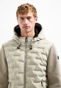 Puffjack in lichtbeige, met een capuchon, ritsluiting, gestructureerde gebreide mouwen en verstelbare trekkoorden.
