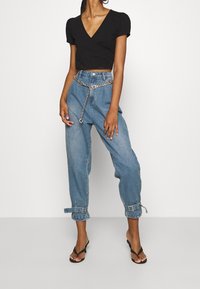 Haut noir portefeuille avec manches courtes bouffantes, associé à un pantalon ample en denim bleu avec revers roulés et une ceinture chaîne argentée. Tongs noires.