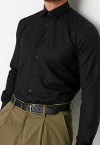 Zwarte lange mouwen shirt met een knoopsluiting kraag, met een gladde textuur en bijpassende knopen, gecombineerd met beige geplooide broek en een zwarte riem.