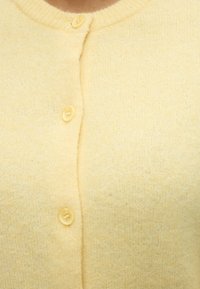 Κοντινή λήψη ανοιχτόχρωμης κίτρινης πλεκτής ζακέτας με τρία κουμπιά που κλείνουν κατά μήκος του μπροστινού μέρους στη μέση.