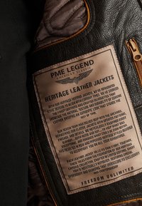 Svart läderjacka med brun etikett som har präglad text. Etiketten beskriver skötsel, designfilosofi och individualitet för varje jacka.