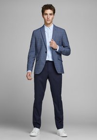 Modra karirana blazer na svetloplavi srajci, v kombinaciji s temnimi hlačami in belimi supergami. Blazer ima žepni ovratnik in en gumb.