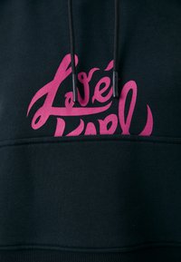 Hanorac negru cu un design mare de text stilizat roz pe față. Include șnururi negre și manșete cu nervuri.