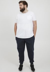 Weißes T-Shirt mit kurzen Ärmeln und Brusttasche, kombiniert mit navyblauen Jogginghosen. Das Modell trägt schwarze Sportschuhe. Neutrale Hintergrund.