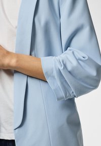 Arm met een lichtblauwe blazer met opgerolde mouwen, gedragen over een wit overhemd en een donkere broek, gezien van opzij ter hoogte van de taille.