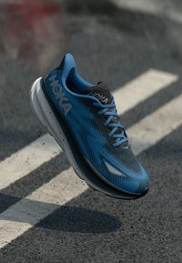 Scarpa da corsa HOKA blu e nera in volo sopra l'asfalto con segnaletica stradale bianca e piccoli detriti volanti.