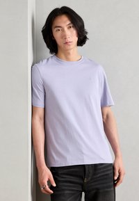 Világos levendulakék crew neck póló puha anyagból, rövid ujjakkal, kis logóval a mellkasán. Sötétszürke farmernadrággal viselve.