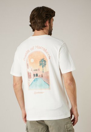 Homme portant un t-shirt blanc avec un motif pastel représentant une piscine, un palmier et des arches sous le texte "Oasis de Marrakech" au dos.