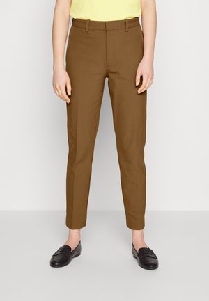 Persoon die bruine op maat gemaakte enkelhoge broek, zwarte loafers en een bleekgele top draagt, staand tegen een effen achtergrond.
