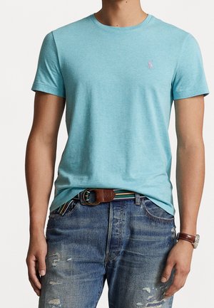 Man wearing a light blue fitted short-sleeve T-shirt tucked into distressed blue jeans with a striped belt and a wristwatch.