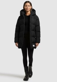 Giacca a vento nera con cappuccio, caratterizzata da un design trapuntato, chiusura lampo frontale e tasche laterali. Indossata sopra un top nero e leggings.