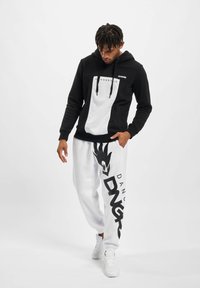 Felpa con cappuccio bianca e nera con logo frontale e pantaloni felpati bianchi caratterizzati da un grande design nero e dal nome del marchio. Sneakers bianche completano il look.