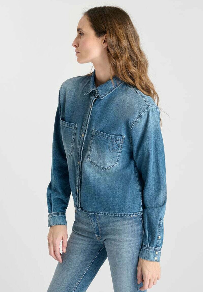 Denim shirt in light blue, featuring a collared neck, two front pockets, and button closure. Long sleeves with buttoned cuffs.