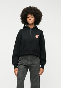Czarna, krótka bluza z kapturem z grafiką motyla na piersi, z regulowanym kapturem i ściągaczami na rękawach. W połączeniu z szarymi dżinsami w wzory.