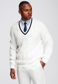 Maglione bianco a trecce con scollatura a V e rifiniture blu navy, caratterizzato da un motivo testurizzato. Indossato sopra una camicia azzurro chiaro e pantaloni coordinati.