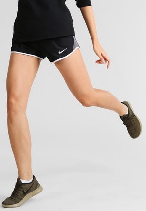 Persoon in zwarte Nike-shorts en olijfgroene hardloopschoenen, halverwege de stap met één been opgetild en één arm gebogen tegen een eenvoudige grijze achtergrond.