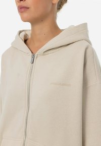 Beige zip-up hoodie med huva, med en jämn textur och en subtil logo på bröstet. Enkelt och avslappnat design.