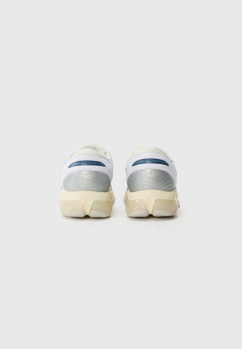 Chaussures de sport blanches avec une semelle grise et beige, présentant un dessus lisse, un col rembourré et un logo bien visible à l'arrière.