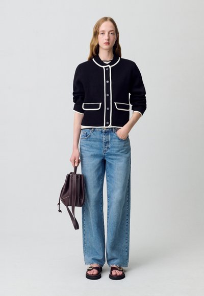 Cardigan cropped nero con bordo bianco, chiusura a bottoni e tasche, abbinato a jeans a gamba larga azzurri e sandali neri, mentre tiene una borsa marrone.