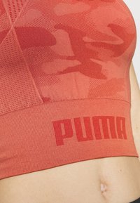 Sports-bh i en strålende koralfarve med et struktureret camouflage-mønster, ribbet bundkant og det dristige "PUMA" logo i rød.