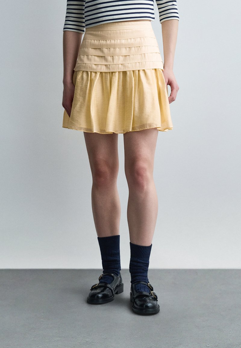 Άτομο που φοράει κίτρινη πιετέ μίνι φούστα, μαύρα μοκασίνια με χρυσά αγκράφες, ναυτικά καλτσάκια και ριγέ λευκό-μαύρο τοπ.