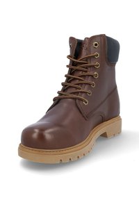 Bota de cuero marrón con cordones, collar negro acolchado y suela de goma beige con tacos, vista frontal en ángulo sobre fondo blanco.