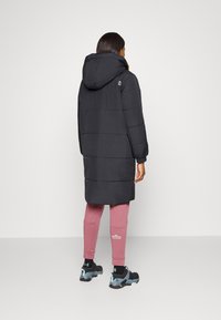 Abrigo largo acolchado negro con capucha, con puños acanalados y detalle de logo. Combinado con pantalones deportivos rosas y zapatillas deportivas negras.