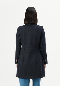 Personne avec des cheveux noirs mi-longs portant un manteau navy ajusté avec des boutons dorés aux manches et un jean bleu, de dos sur fond blanc.