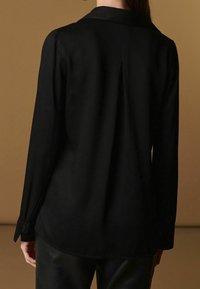 Osoba ve dlouhém černém tričku s plisováním na zádech a polo límcem, v kombinaci s černými kalhotami, na jednobarevném hnědém pozadí.