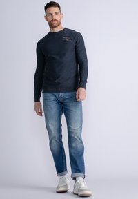 Granatowy sweatshirt z długimi rękawami i okrągłym dekoltem, z logo na piersi, zestawiony z jasno niebieskimi dżinsami i białymi sneakersami.