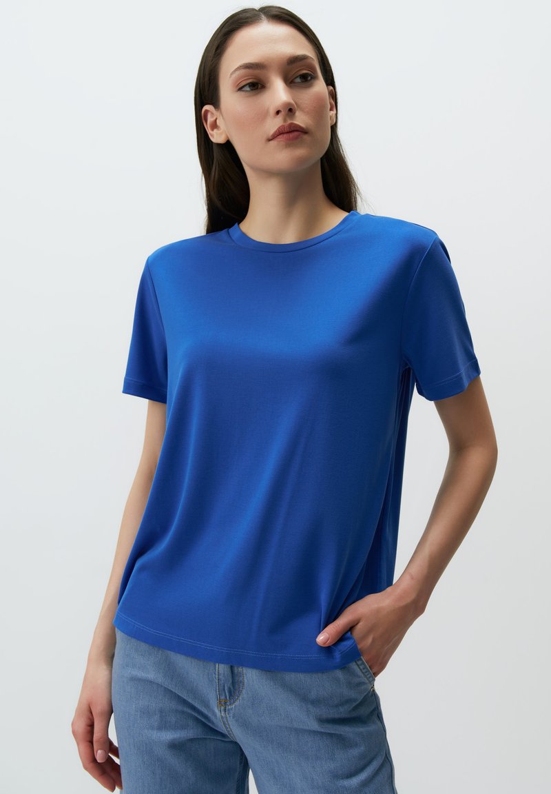 Niebieska koszulka z krótkim rękawem, okrągłym dekoltem, wykonana z gładkiego materiału, o luźnym kroju z detalami wzdłuż bocznych szwów, zestawiona z dżinsami.