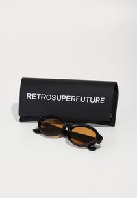 Mustad ovaerilised prillid pruunide läätsedega, kaasas matt must isase, millel on valge kiri "RETROSUPERFUTURE."