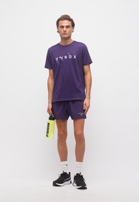 Άνδρας με μωβ αθλητική ενδυμασία Puma HYROX κρατάει ένα κίτρινο μπουκάλι νερού Puma, φορώντας λευκές κάλτσες και μαύρα αθλητικά παπούτσια Puma, πάνω σε λευκό φόντο.