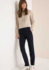 Beige hoodie met trekkoorden, gecombineerd met marineblauwe sportbroek met zijzakken. Afgewerkt met witte sneakers en subtiele merkaanduidingen.