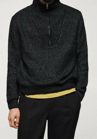 Schwarzer Strickpullover mit hohem Kragen und halbem Reißverschluss, der aus strukturiertem Material gefertigt ist und über einem gelben Hemd einen Lagenlook präsentiert.
