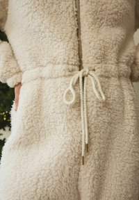 Cremefarbener Fleece-Overall mit strukturiertem Obermaterial, goldfarbenem Reißverschluss und einer Taillenschnürung mit Kordelenden und Metallelementen.